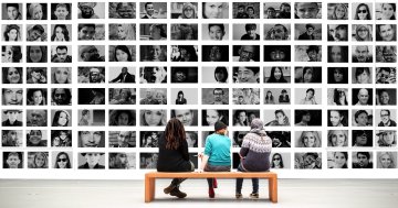 3 Jugendliche sitzen vor einer riesigen Fotowand. Die Fotos zeigen unterschiedliche Menschen.